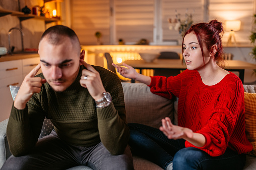 A man and woman are seated on a couch together, arguing. The man is plugging his ears with his fingers. The woman is trying to talk to him and has her hands out beside her.