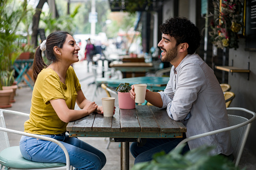 A Latin American couple is seen on a coffee date at an outdoors coffee shop. They are sitting across from one another, both holding cups of coffee and smiling at each other. They are in conversation with one another and are enjoying each others company.