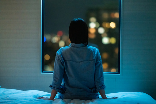 In this image, a woman is seated on her bed looking out her bedroom window. It is night time, and outside the window you can see the night sky and the city lights.