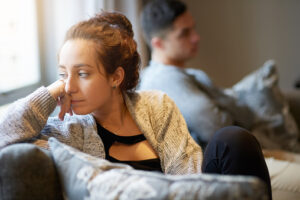 A young couple is sitting next to each other on their living room couch. They are upset with one another after having an argument, as they both are looking away from each other. The woman has a sad facial expression as she leans her head on her hand.