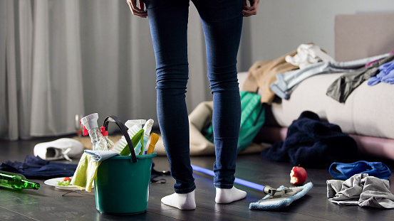A woman is standing in her home surrounded by a mess. There is a bucket next to her feet filled with cleaning supplies. On the floor there are clothes and dirty dishes.