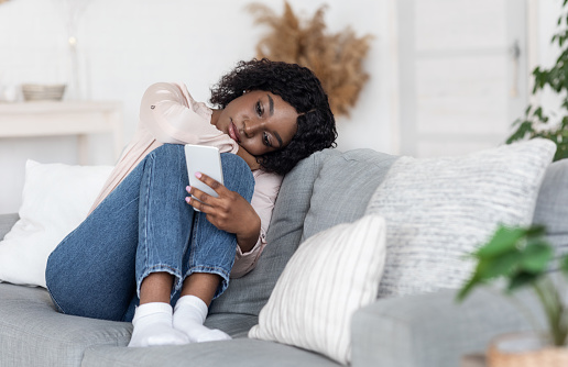 A young African American woman is sitting on the couch with her legs propped up. She has a sad facial expression as she looks at her phone in her hand.