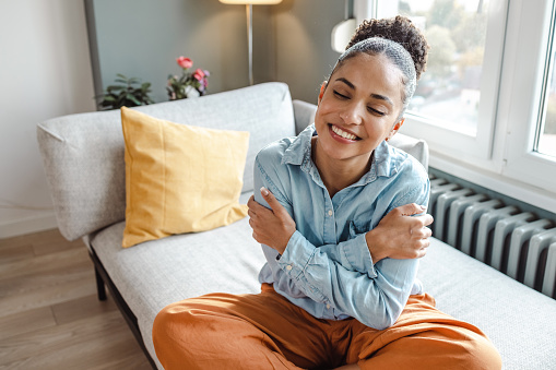 A young African American woman is sitting in her home on a sunny morning. She is smiling while her eyes are closed and she is hugging herself.