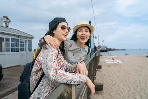 Two young Asian women are spending time together on the pier of a beach. They are standing close together as one puts her arm over the other. They are both happy and enjoying their time at the beach.