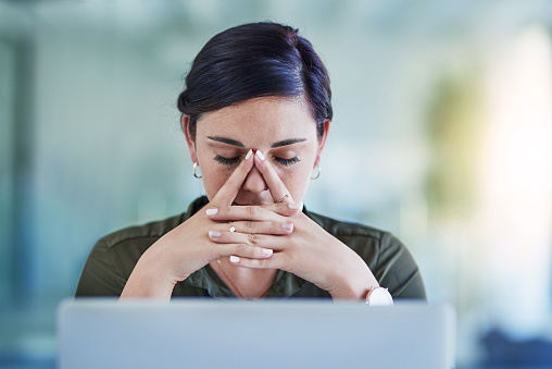 A young businesswoman is sitting her her office with her hands on her face. She has her laptop in front of her and has a stressed out facial expression.