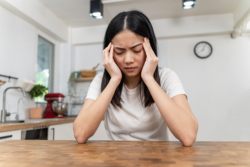 A young Asian woman is sitting in her kitchen at the dining table. She is holding her hands to her temples to relieve her headache as she closes her eyes.