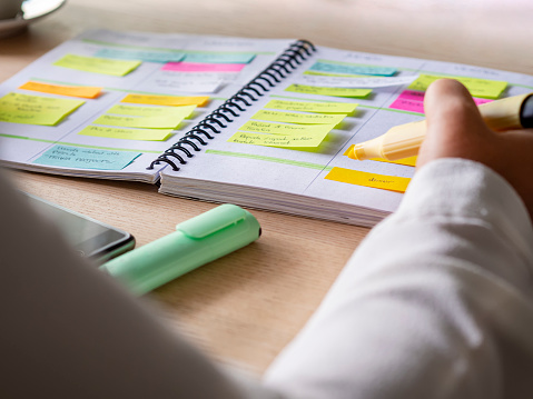 In this photo we see a close-up of an agenda organized with color-coding sticky notes for time management. The hand is holding a yellow highlighter marker. 