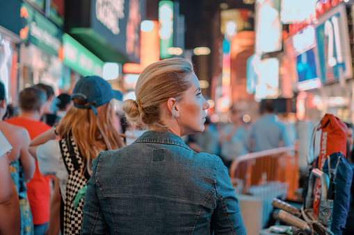 A young blonde woman is seen looking around while walking in New York, at Times Square. Her blonde hair is tied in a bun as she is looking to her side.