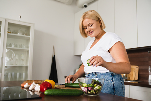 A beautiful plus sized blonde woman is standing in her kitchen preparing a meal for herself. She has vegetables set on the counter in front of her and is making a salad.