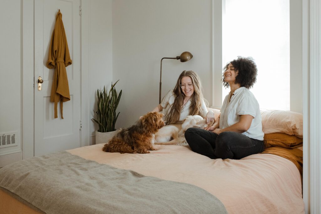 A young couple is sitting on their bed with their 2 dogs. The two women are both laughing and smiling as they pet their dogs and enjoy each other's company.