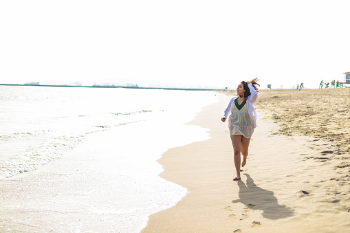 A young woman is running on the shore of the beach on a beautiful sunny morning. The waves are calm as the woman runs alongside them.
