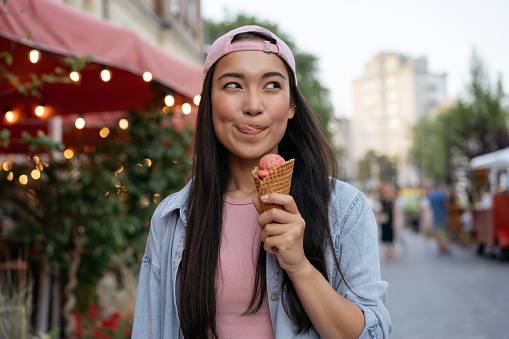 A young woman is outside holding an ice cream in her hand. She is licking her lips and smiling as she enjoys her ice cream. She is wearing a pink hat backwards.