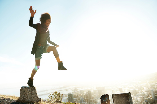 A young black woman is outdoors jumping from pillar to pillar on the edge of a mountain road. The sun is shining and the view of the city is behind her. She is smiling as she is leaping to the pillar in front of her.