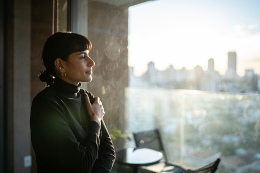 A young woman is standing in her apartment, looking out to the city view. She has a content look on her face as she looks out the window.