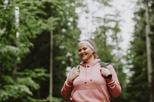 A young woman is taking a walk in the forest with her backpack on her. She is smiling as she walks in nature.