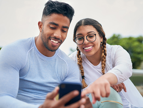 A young couple is enjoying themselves in the outdoors. They are both sitting on the grass as they look at pictures on the mans phone.