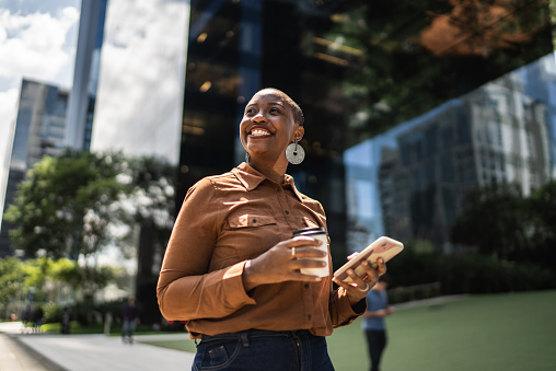 A black woman is standing outside in front of an office building, holding a coffee cup in one hand and her phone in the other hand. She is smiling and looking around.