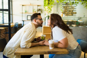 A couple is sitting at a cafe across from each other. They are both leaning forward on the table, going in for a kiss. They are holding each others hands across the table.