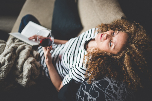 A young curly haired woman is laying down on her couch relaxing. She has an open book in her lap, and a glass of red wine in her hand.