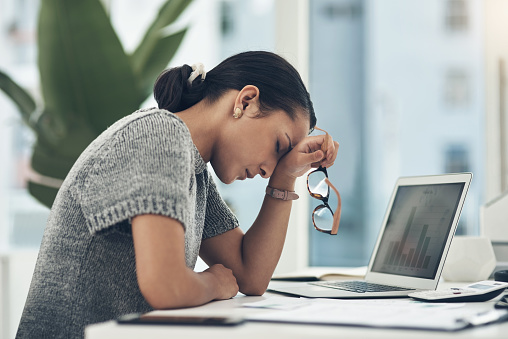 A working woman is sitting at her office, stressed out and tired from working. In front of her is her laptop displaying a chart. She has her head resting on her hand with her seeing glasses in her hand. Her eyes are closed as she takes a minute for herself.