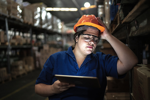 A young woman is at a warehouse working as a constructor. She is wearing a hard hat and protective glasses. She is holding a tablet in her hand, and is resting her head as she closes her eyes.
