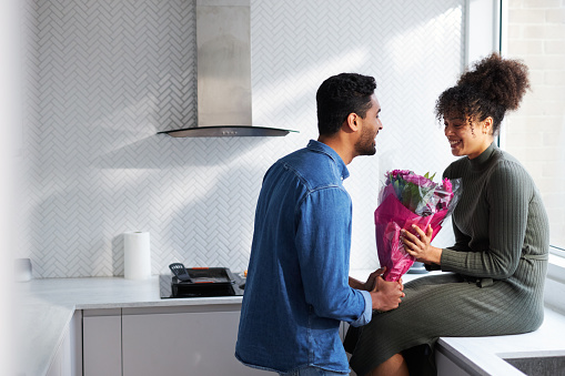 A young happy couple is together in their kitchen. The woman is sitting on top of the counter as the man gives her a beautiful pink flower bouquet. 