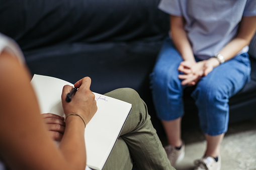 In this photo, we see a psychotherapy session. A woman is talking to her therapist as she sits on a black couch. The therapist is writing chart notes in her notebook as the client talks to her.