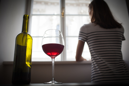 A sad woman is looking out of her window with her back facing the camera. She is wearing a striped short sleeve shirt. Closer to the frame is a bottle of wine and a glass of red wine next to it.