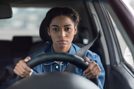 A young African American woman is driving a car, sitting in traffic. Both of her hands are on the steering wheel as she is gazing in front of her. 