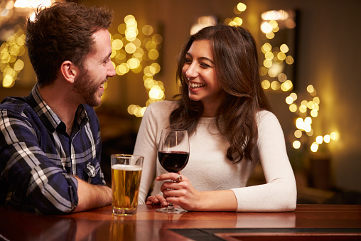 A young couple is on a date together. They are sitting next to each other and smiling. The woman is holding a glass with red wine in it. The man has a beer in front of him on the table.