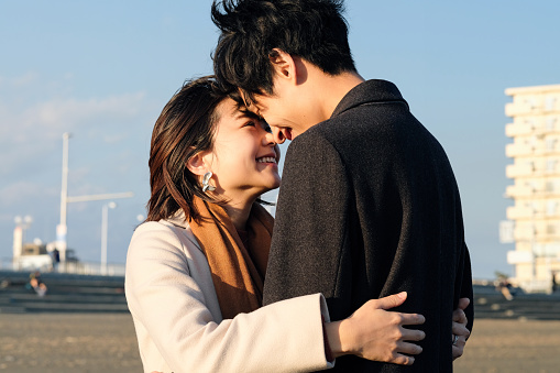 A young Asian couple is embracing each other at the beach. They are hugging one another and looking in each others eyes as they smile.