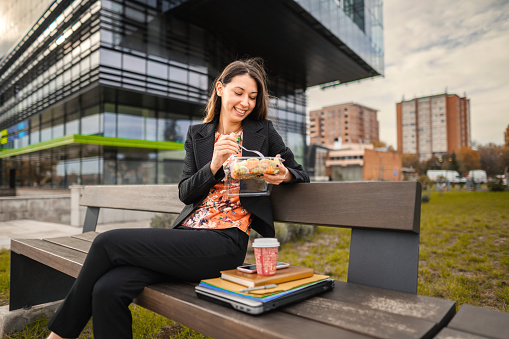 A young business woman is sitting in front of her office building having lunch. She is sitting on a bench with her laptop and notebook beside her. Her coffee is placed on her notebook next to her. She is eating a salad.