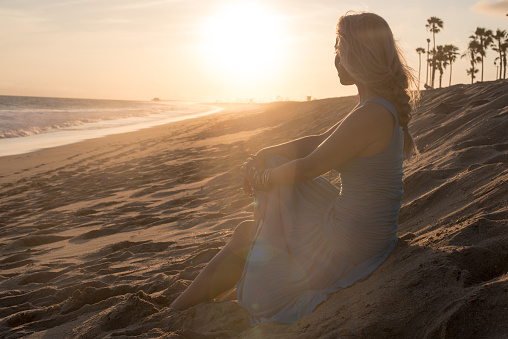 A young blonde woman is sitting on the sand at the beach enjoying the sunset and waves. The sun is shining on her.