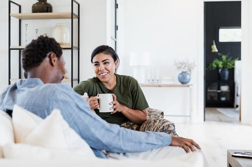 An African American couple is sitting together on their couch in the living room. She is holding a coffee mug as they engage in conversation and smile at each other.