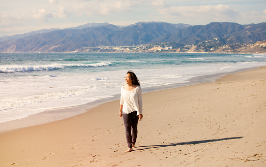 A young woman is enjoying a peaceful walk on the shore of a sunny beach. She is enjoying the view of the waves and the breeze of the ocean.