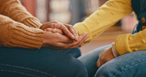In this photo there are two people holding hands and getting together in unity. The woman on the left is wearing an orange sweater, and the woman on the right is wearing a yellow long sleeve sweater.