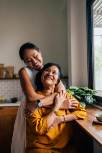In this photo we see an Asian mother and daughter sitting in their kitchen. The mother is sitting on a chair, as her daughter stands behind her and hugs her. They are smiling at one another and embracing each other.