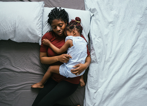 In this photo there is an African American mother laying down in a bed as her young daughter lays on top of her. They are taking a nap together in the bedroom.