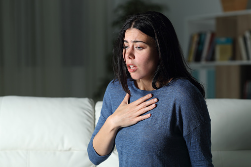 In this photo there is a young woman in her home at night. She is suffering from an anxiety attack as she puts her hand on her chest and has a distressed facial expression.