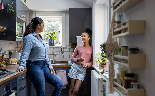 An African American mother and daughter are standing together in the kitchen and talking. The mother is leaning on the kitchen counter and the daughter is leaning on the kitchen wall.