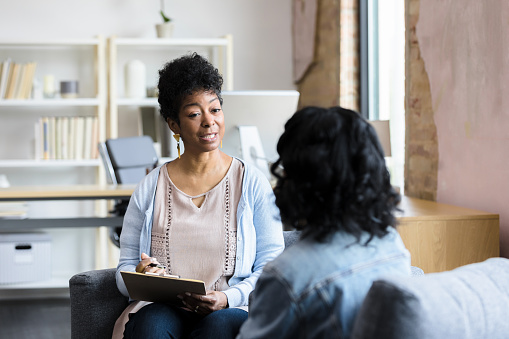 An African American woman therapist is sitting across from her client in her workspace. She is holding up a note chart as she speaks to the client and takes notes.