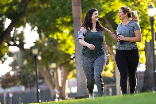 Two young women are out on a jog together in the park. They are running side by side, as one of the women puts her hand on the other woman.