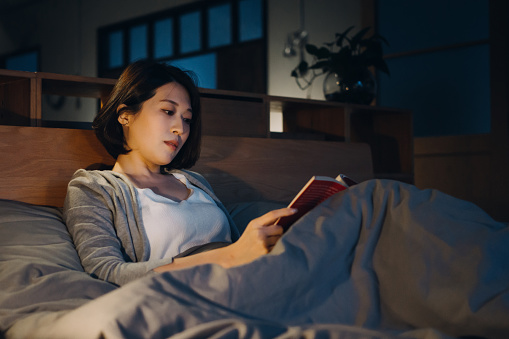 A young Asian woman is laying in bed reading a book at night. She is relaxing and winding down from her day.