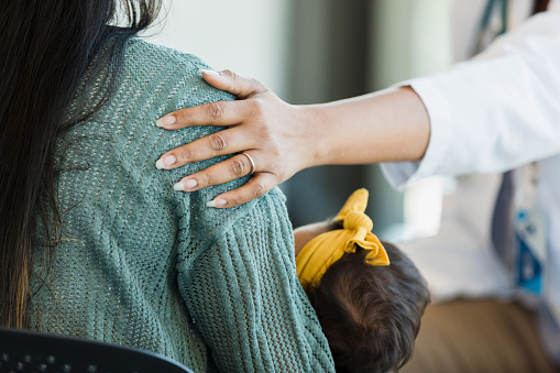 In this photo, an unrecognizable person places their hand on a mother's shoulder as she offers her comfort and support. The mother is holding her infant in her arms, and the infant has a yellow headband on her head.
