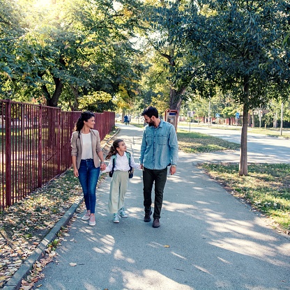In this photo, a mother and father are walking their daughter school. They are walking outdoors on a sunny day, as the daughter walks in the middle of both parents, hand in hand.