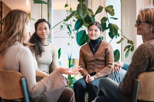 Four women are sitting in a circle participating in a group therapy meeting. Three of the women are looking at the woman on the left corner and listening to her speak. Mental health awareness month. 