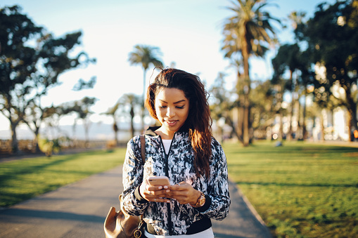In this photo there is a multiracial Latina woman walking outdoors as the sun shines on her. She is looking down at her phone and smiling as she walks.
Mental health awareness month