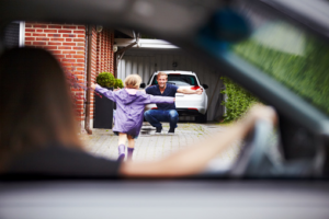 In this photo a mother is sitting in a car looking out the window as she watches her young child run towards the father.