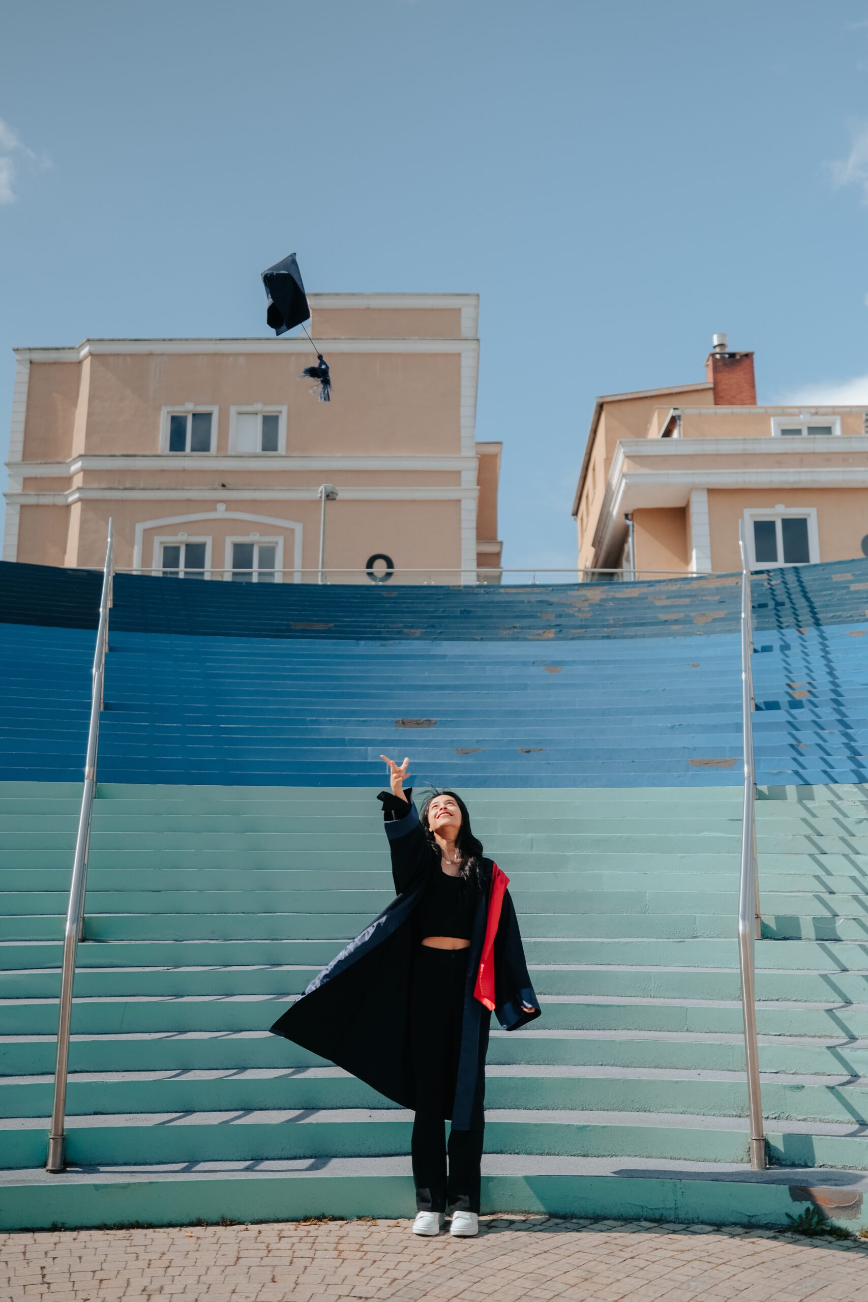 A young woman is standing by a set of blue stairs wearing a black graduation gown with a red sash. She is throwing her graduation cap in the air as she smiles.