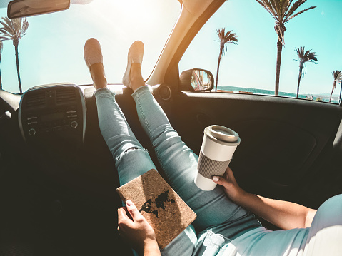 In this photo, a woman is sitting inside of her car with her feet on the dashboard. The car is parked at the beach, as the woman holds a coffee in her hand and a notebook in her other hand.